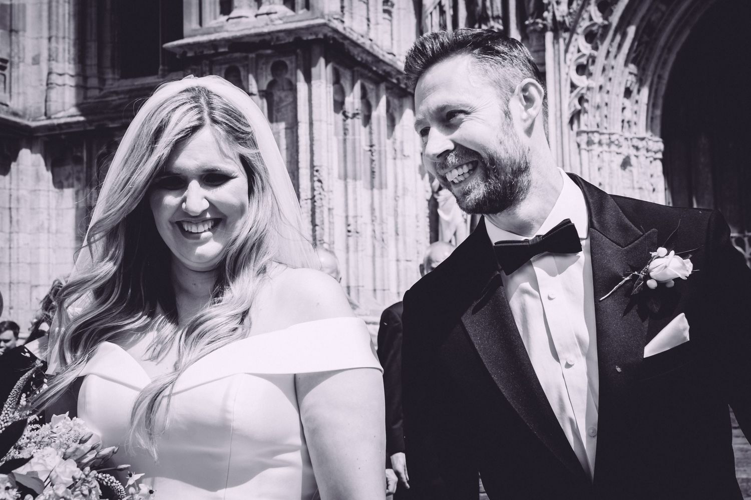 Black and white photo of a smiling bride and groom outside a gothic cathedral on their wedding day.