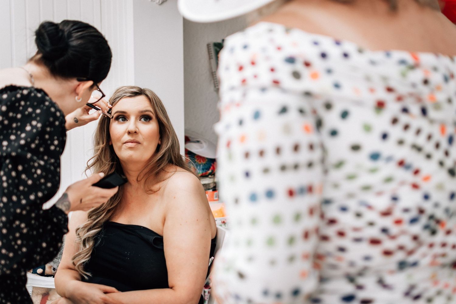 Bridesmaid getting makeup applied by artist while seated during wedding preparation indoors.