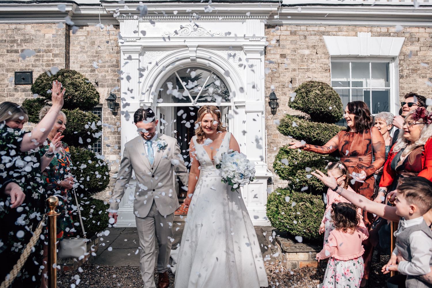 Bride and groom exit venue through confetti shower, surrounded by cheering guests outside elegant white building.