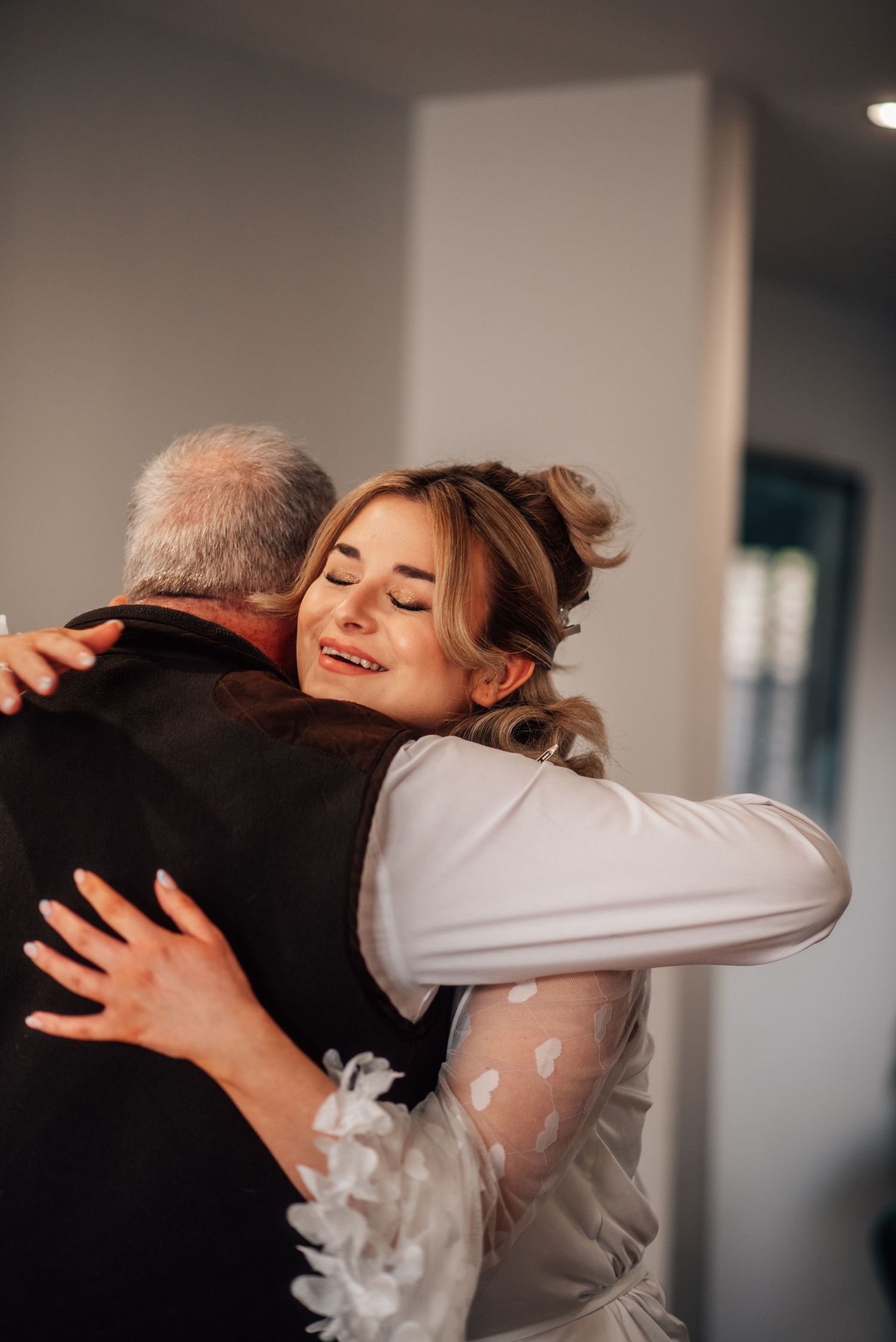 A bride in a white lace dress shares an emotional hug with an older man, smiling with eyes closed.