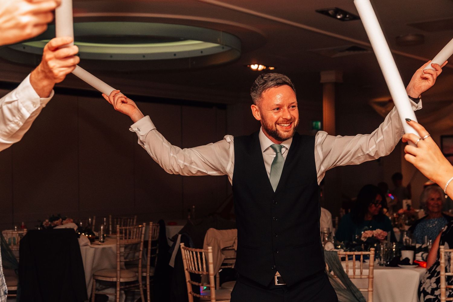 Groom in navy waistcoat celebrates joyfully with raised arms during lively wedding reception dance.