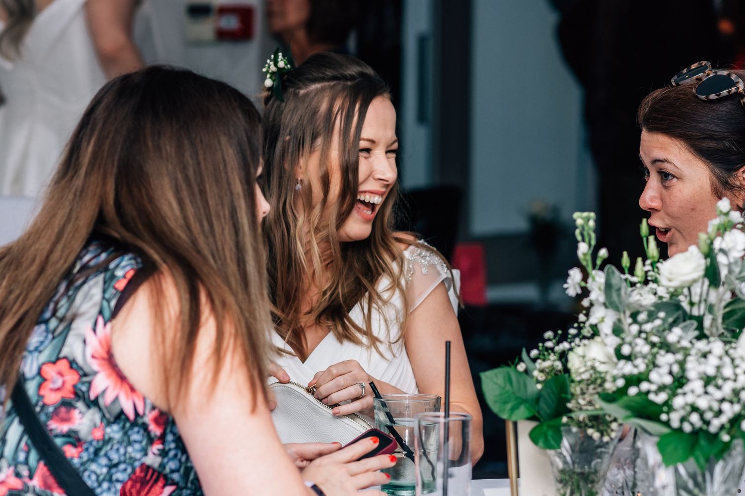 Bride in white dress laughs joyfully with guests at wedding reception table with flowers.