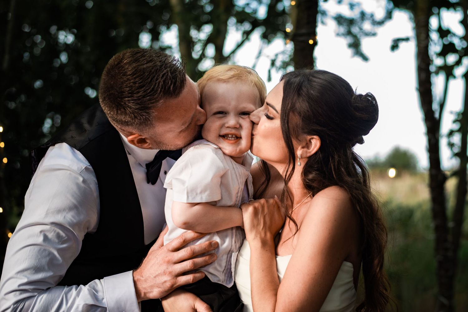Bride and groom in wedding attire kissing a smiling baby on the cheeks outdoors with string lights.