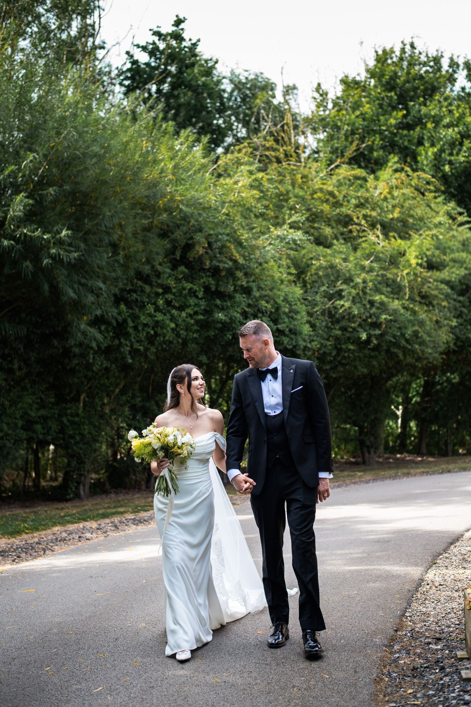 Bride and groom walk hand-in-hand on a path surrounded by lush green trees on their wedding day.