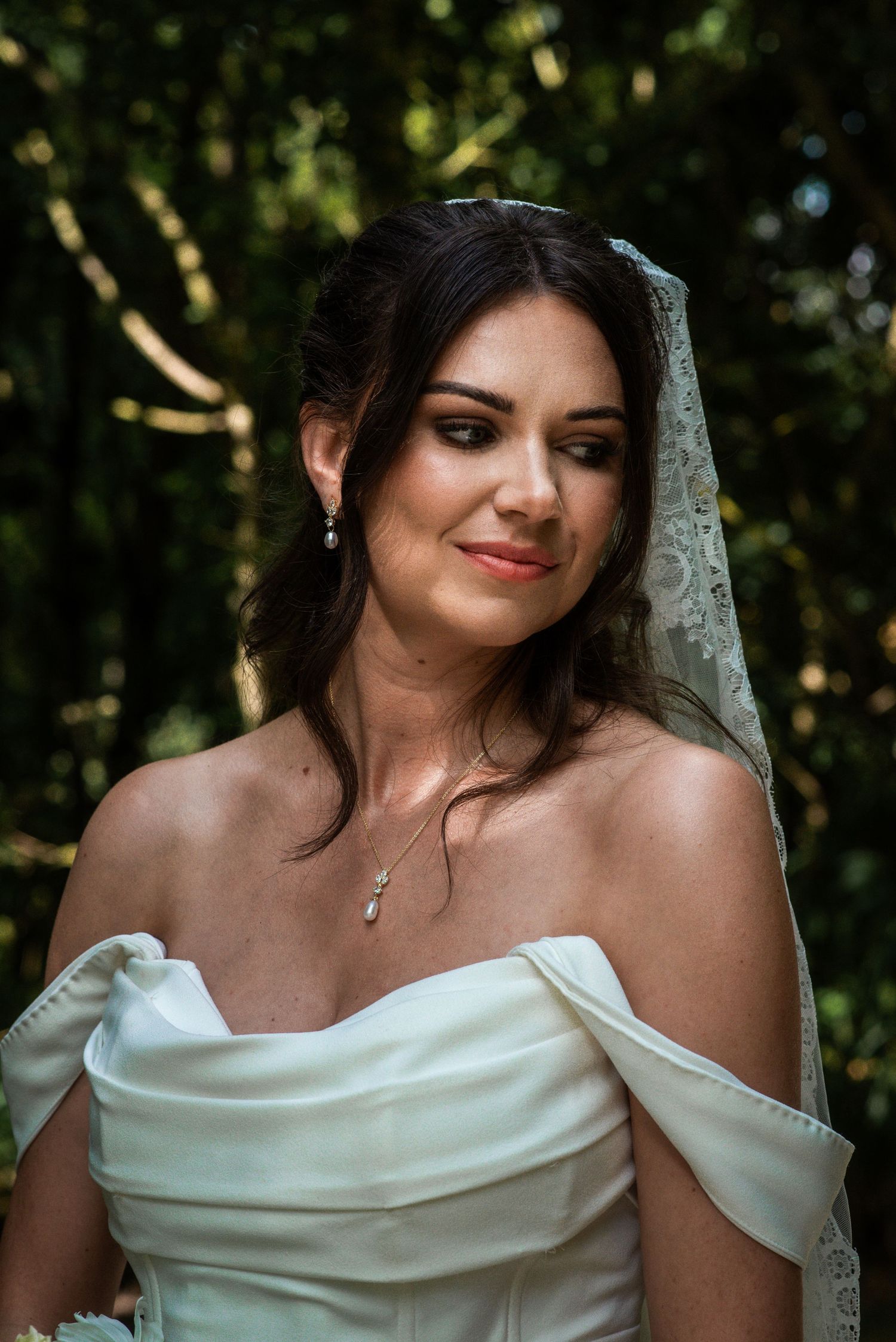 Elegant bride in off-shoulder white gown and lace veil smiling softly outdoors among green trees.