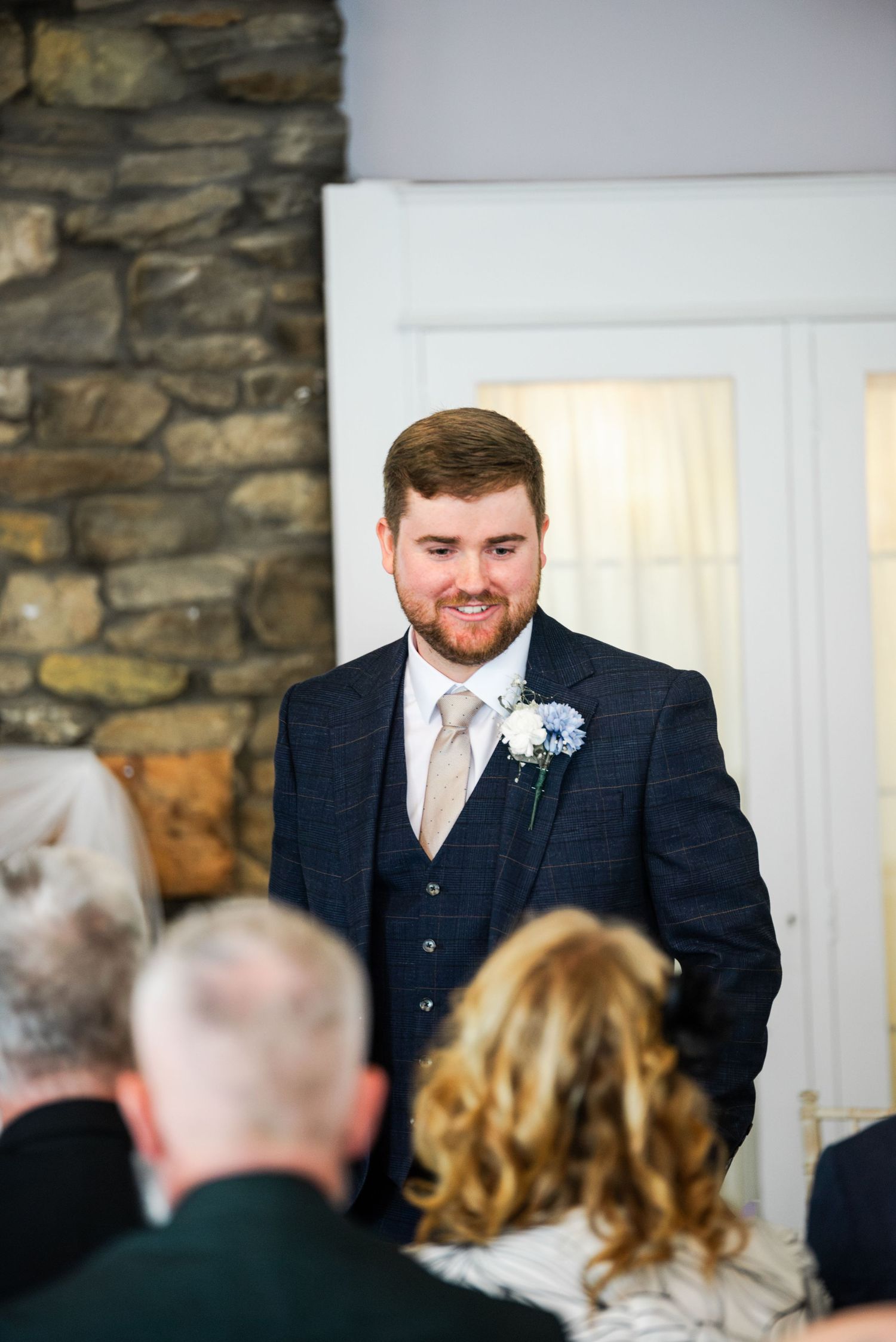 Smiling groom in navy checkered suit with floral boutonniere stands at wedding ceremony altar before seated guests.