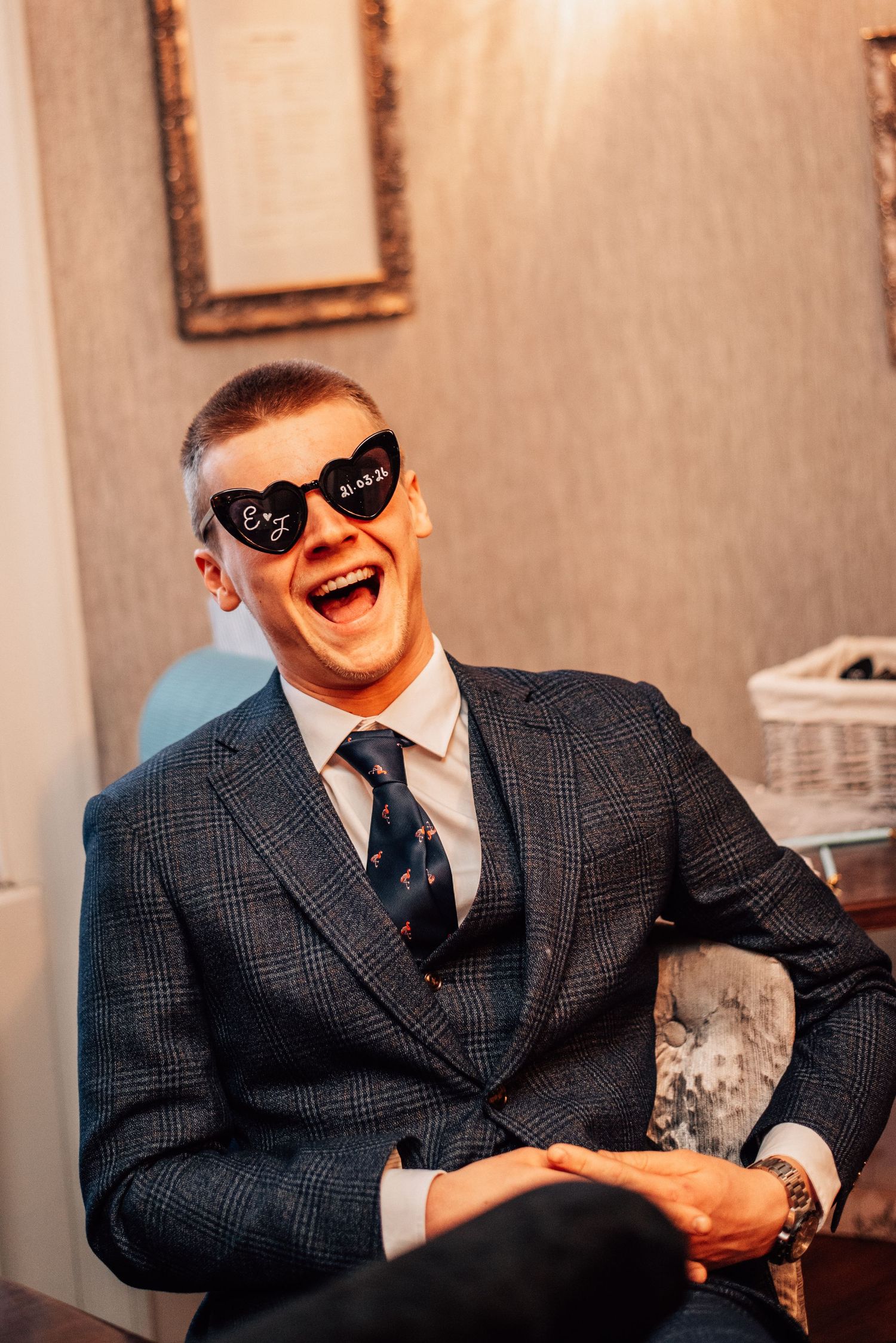 Laughing wedding guest wearing novelty heart-shaped sunglasses in grey checked suit at reception.