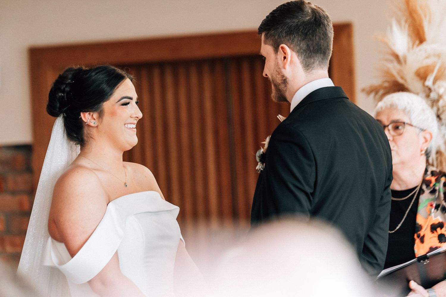 Bride in off-shoulder white gown smiling at groom during wedding ceremony with officiant in background.