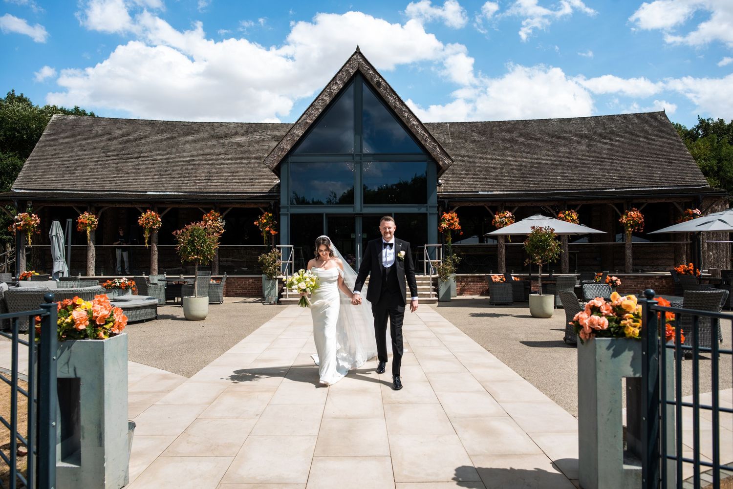 Bride and groom walk hand-in-hand outside a rustic barn wedding venue on a sunny day with orange floral decor.