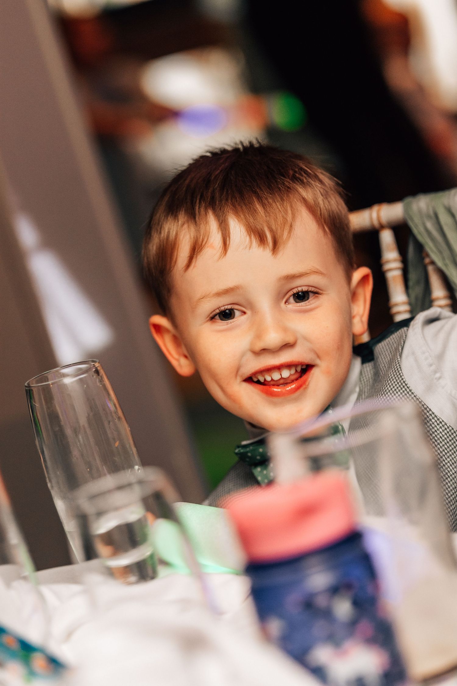 A smiling young boy with short brown hair sits at a table with glasses, looking cheerful and happy.
