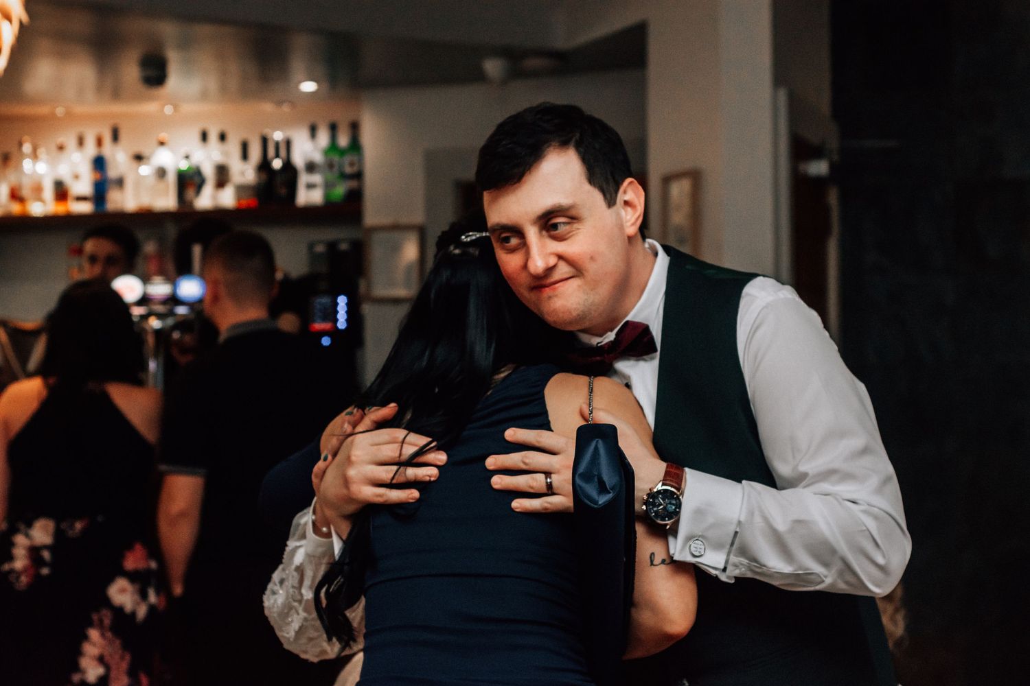A man in a vest and bow tie hugs a woman in a blue dress at a bar during a wedding reception.