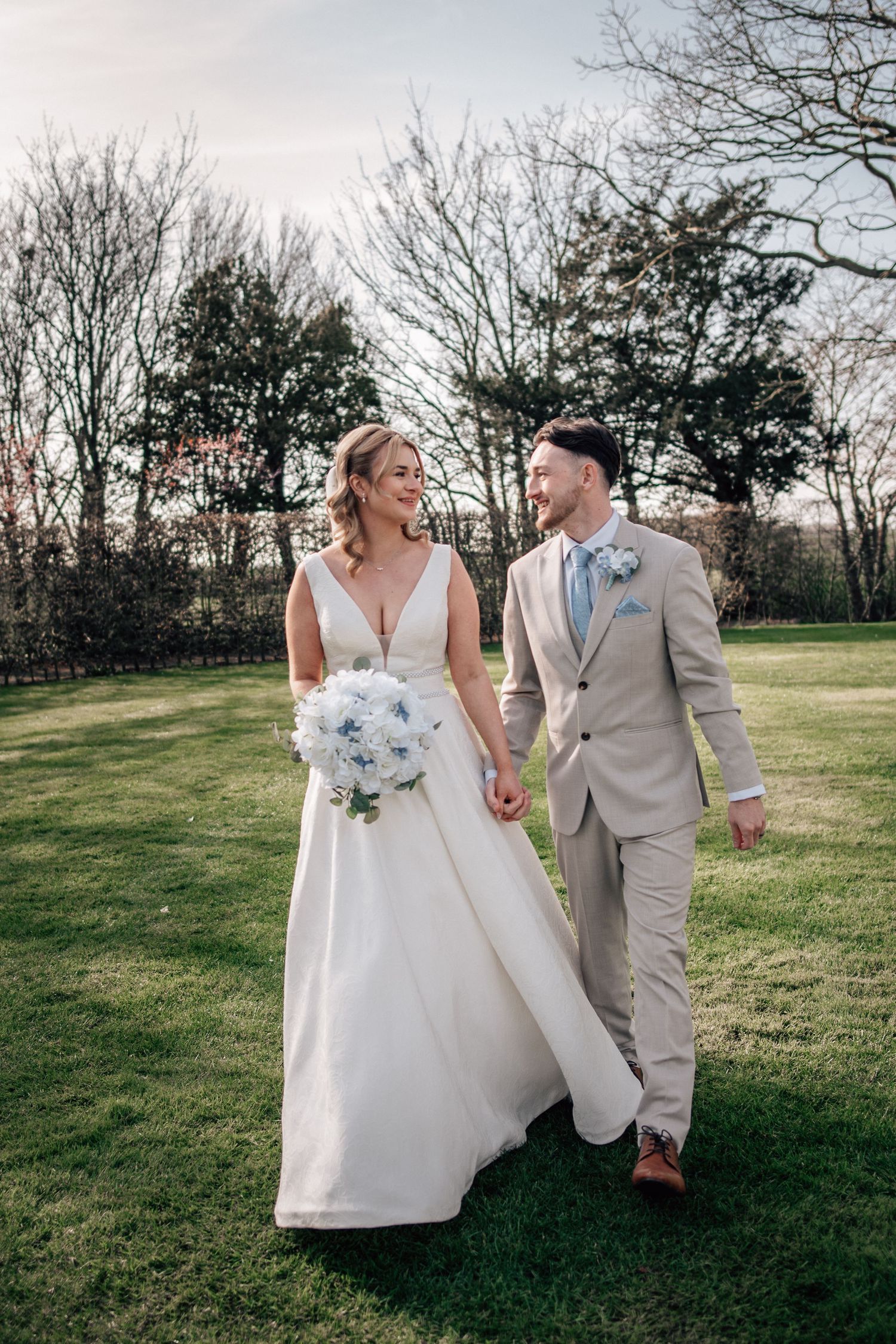 Bride and groom walk hand-in-hand on green lawn, she holds blue bouquet, he wears tan suit with floral boutonniere.