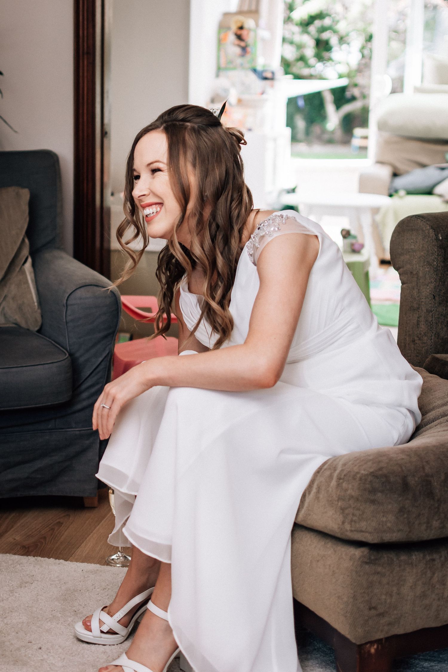 Smiling bride in white lace dress seated in armchair during wedding morning preparations at home.