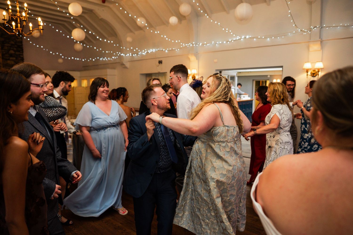Wedding guests dancing and celebrating joyfully in a decorated venue with fairy lights and paper lanterns.