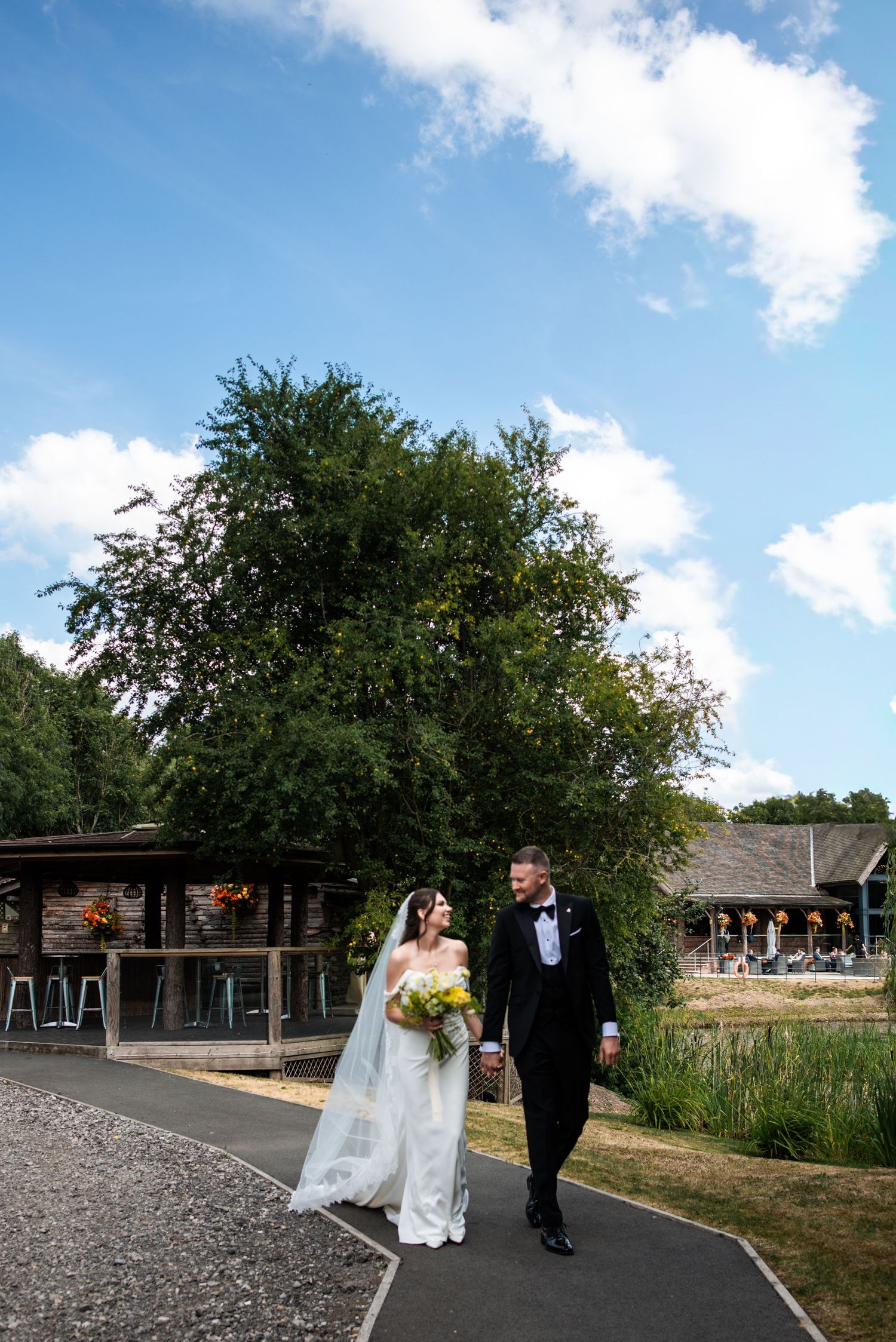 Bride and groom walk hand-in-hand on a sunny day, bride in white gown with veil, groom in black tuxedo.