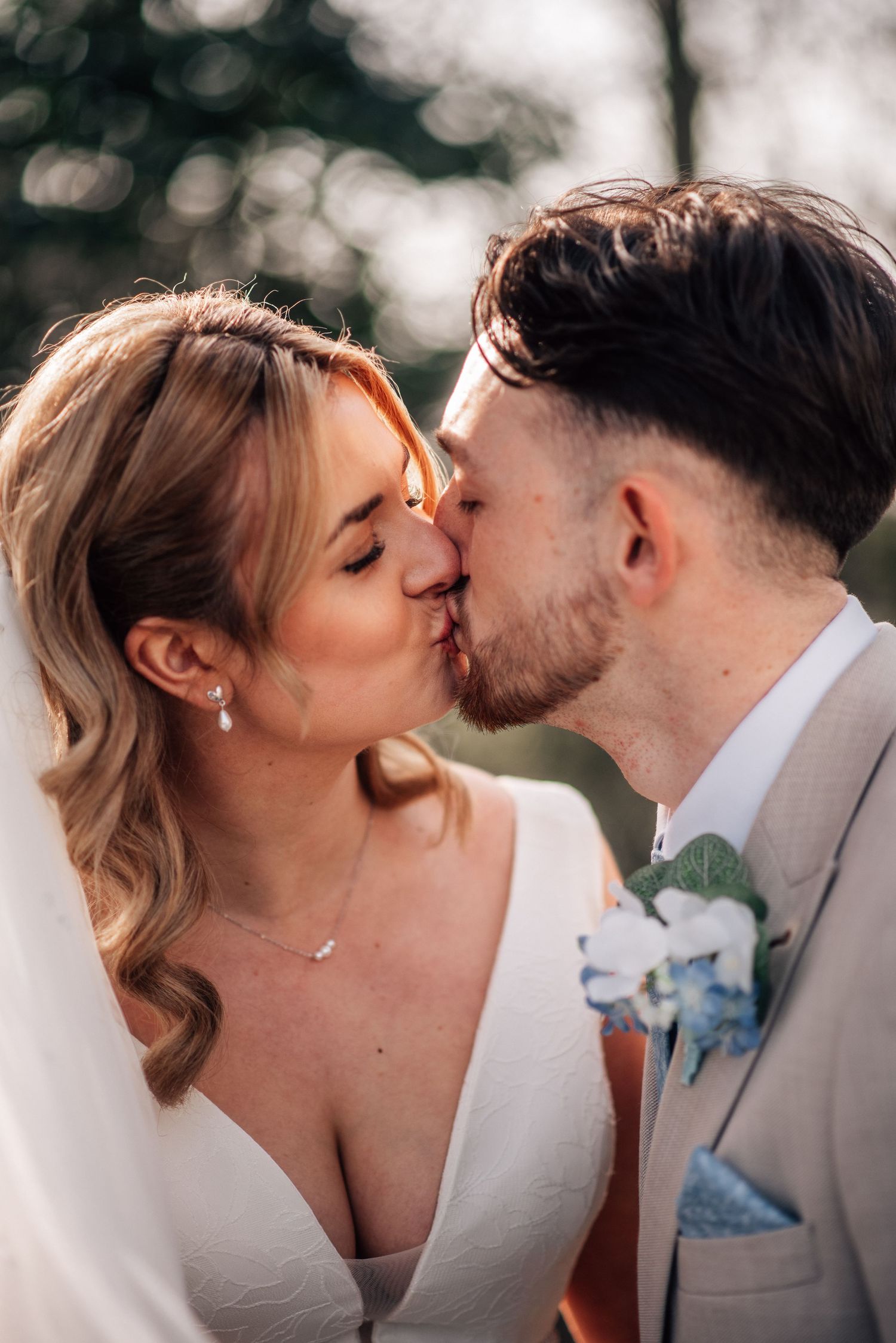A bride and groom share a romantic kiss outdoors on their wedding day, with soft bokeh background lighting.