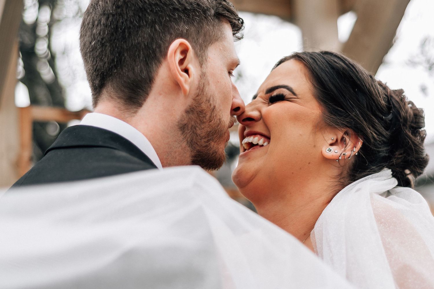 A joyful bride laughs as her groom leans in for a kiss, with her veil flowing in the foreground.