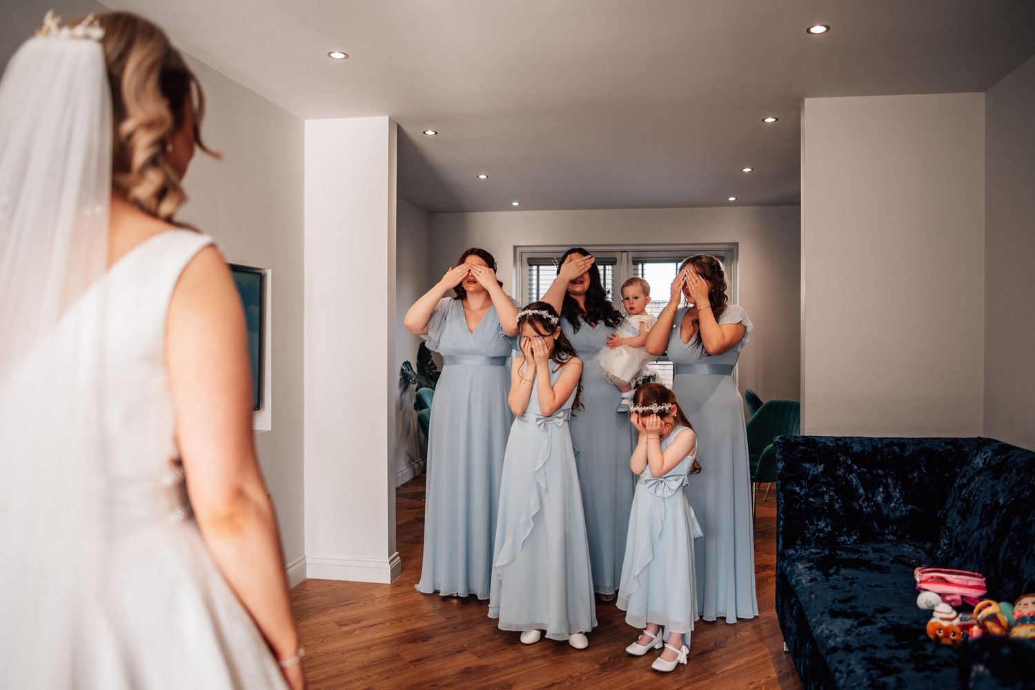 Bridesmaids and flower girls react with joy as bride reveals her wedding dress.