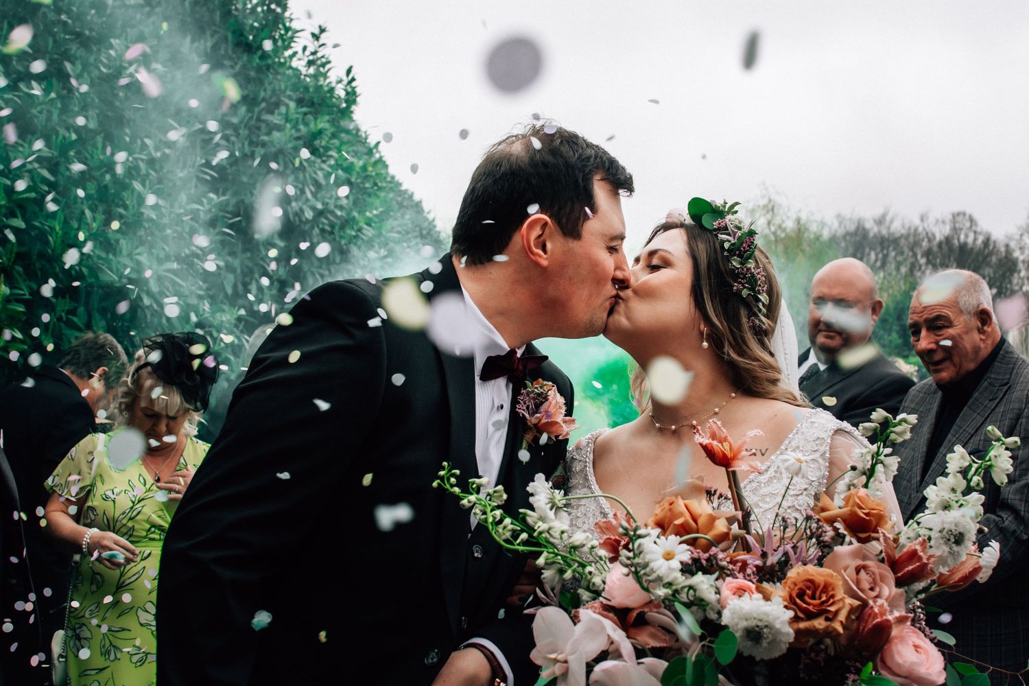 Bride and groom share a kiss surrounded by white confetti, bride holding colorful bouquet at outdoor wedding ceremony.