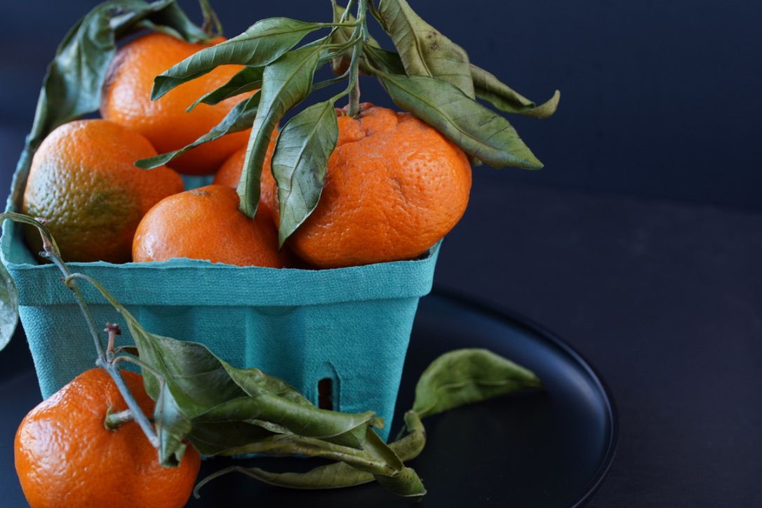 Fresh mandarin oranges with green leaves in turquoise basket against dark background.