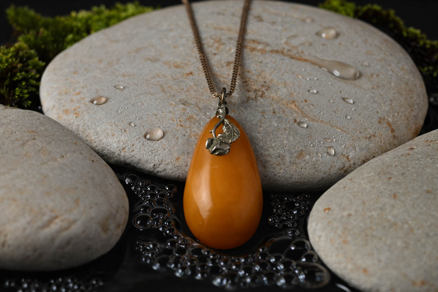 Amber teardrop pendant necklace on leather cord displayed on smooth gray stones with water droplets.