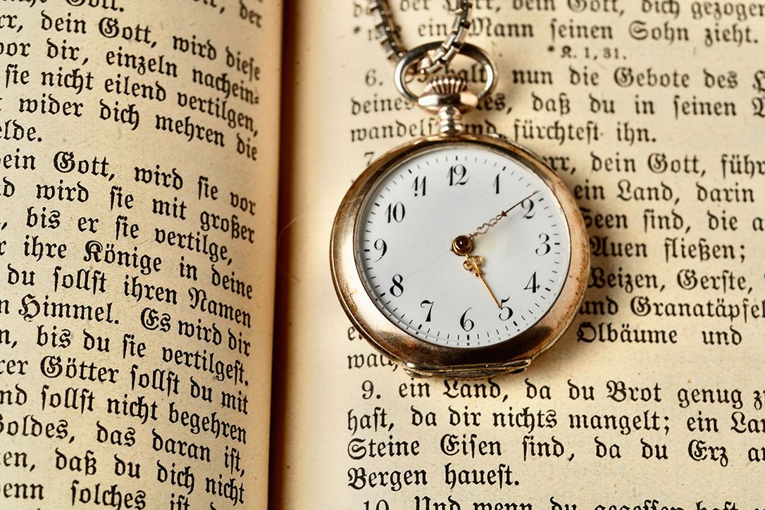 A vintage pocket watch sits open atop an antique book with German text visible on its yellowed pages.
