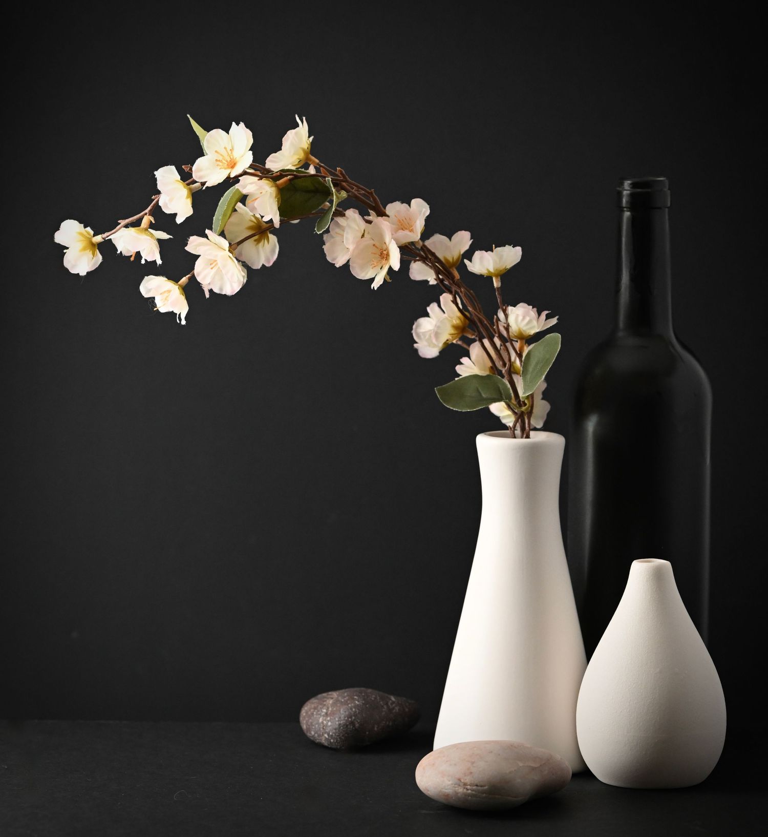 Cherry blossoms in white ceramic vases with wine bottles against dark background.