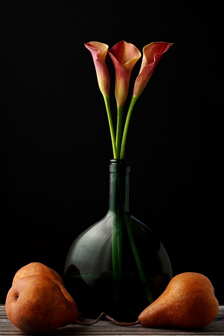 Orange calla lilies in vase with sweet potatoes arranged on wooden surface.