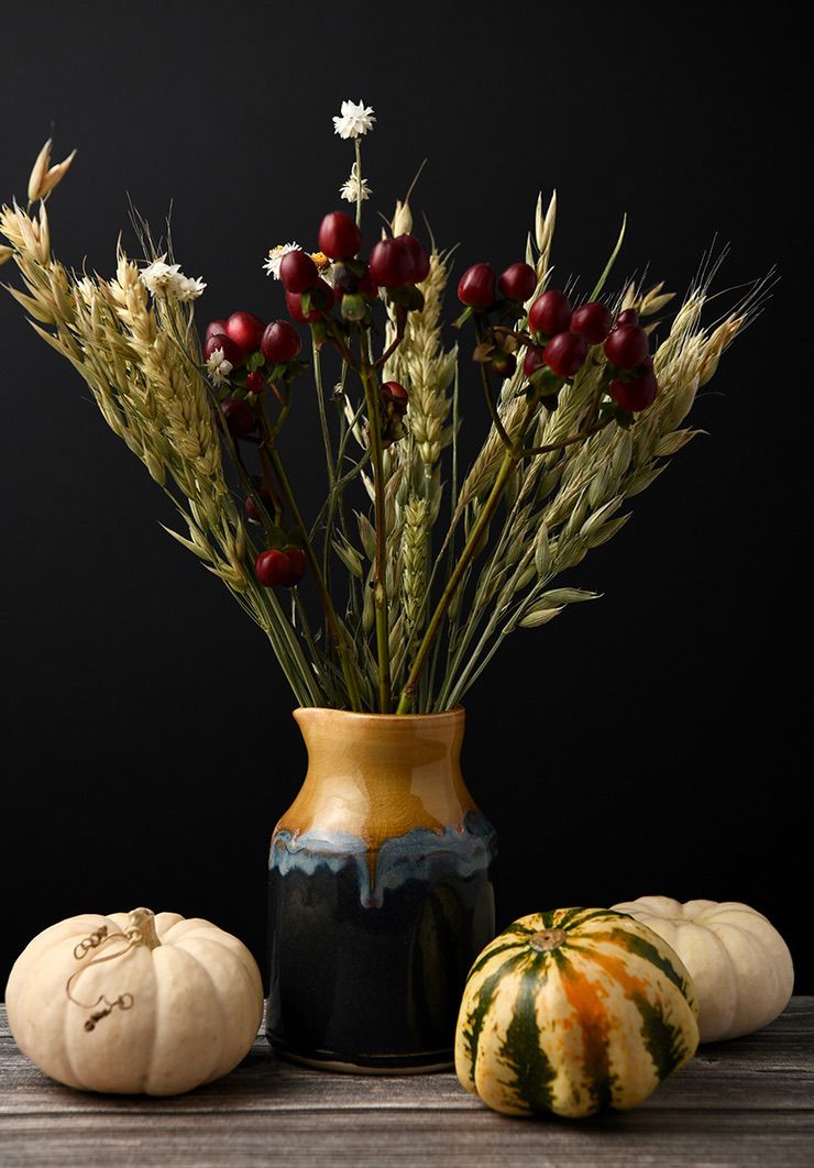 Rustic arrangement of wheat stalks and red berries in two-toned vase with decorative pumpkins.