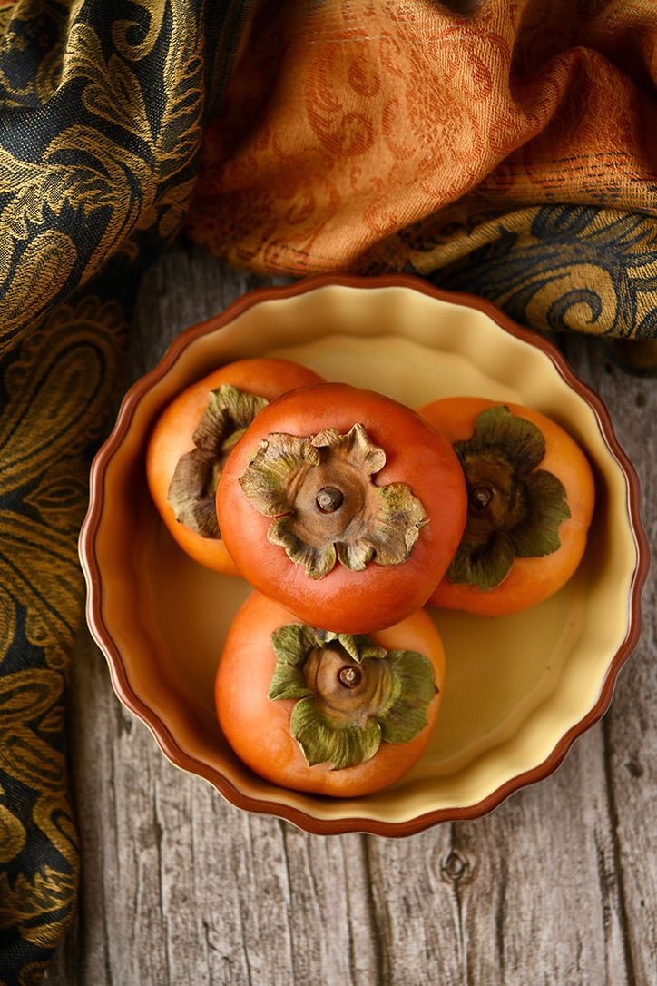 Three ripe persimmons displayed in a vintage orange ceramic bowl on a rustic wooden surface.