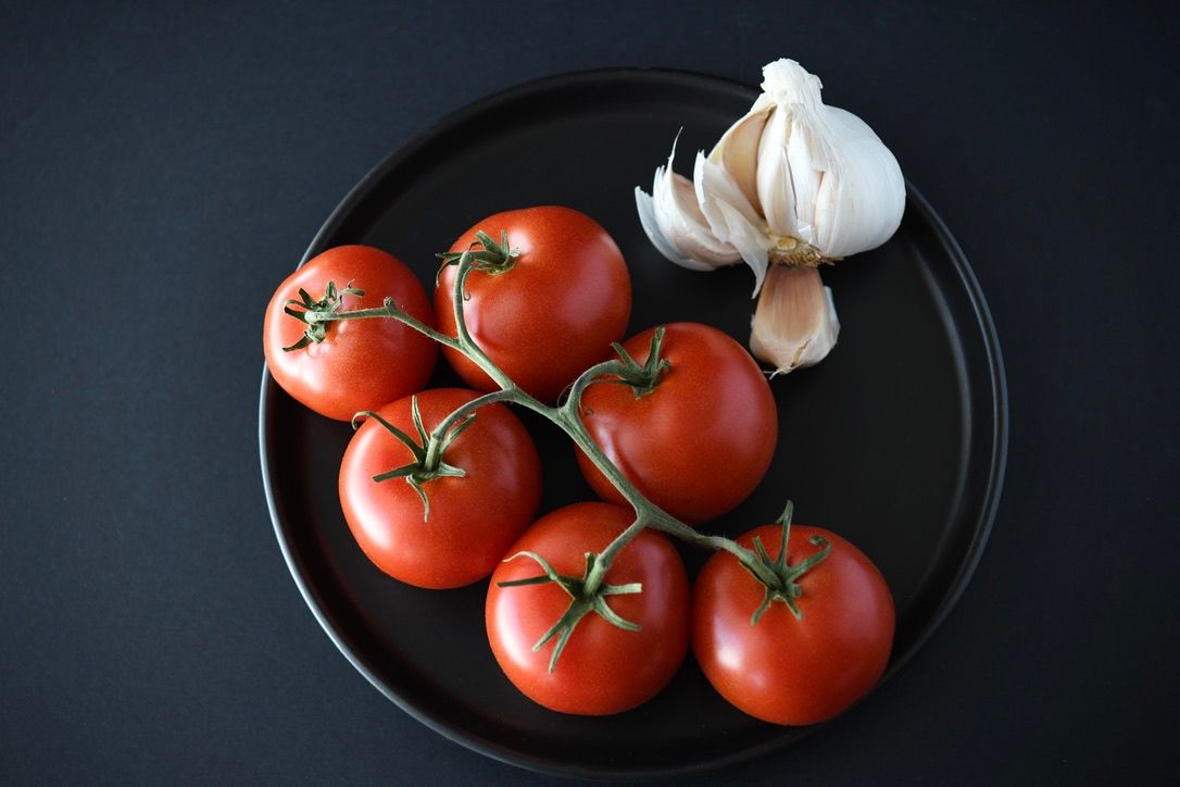 Fresh tomatoes and garlic arranged on black plate against dark background.