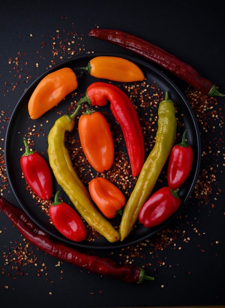 Colorful sweet peppers and chili peppers arranged on black plates with scattered red spices on dark background.