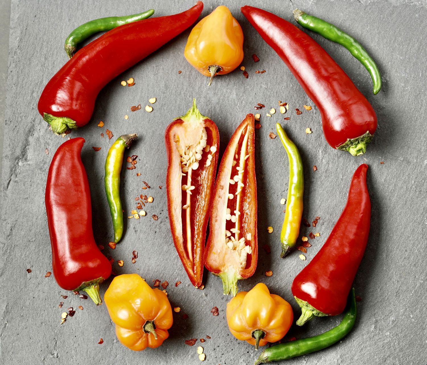 Variety of colorful chili peppers arranged on grey stone surface with scattered seeds.