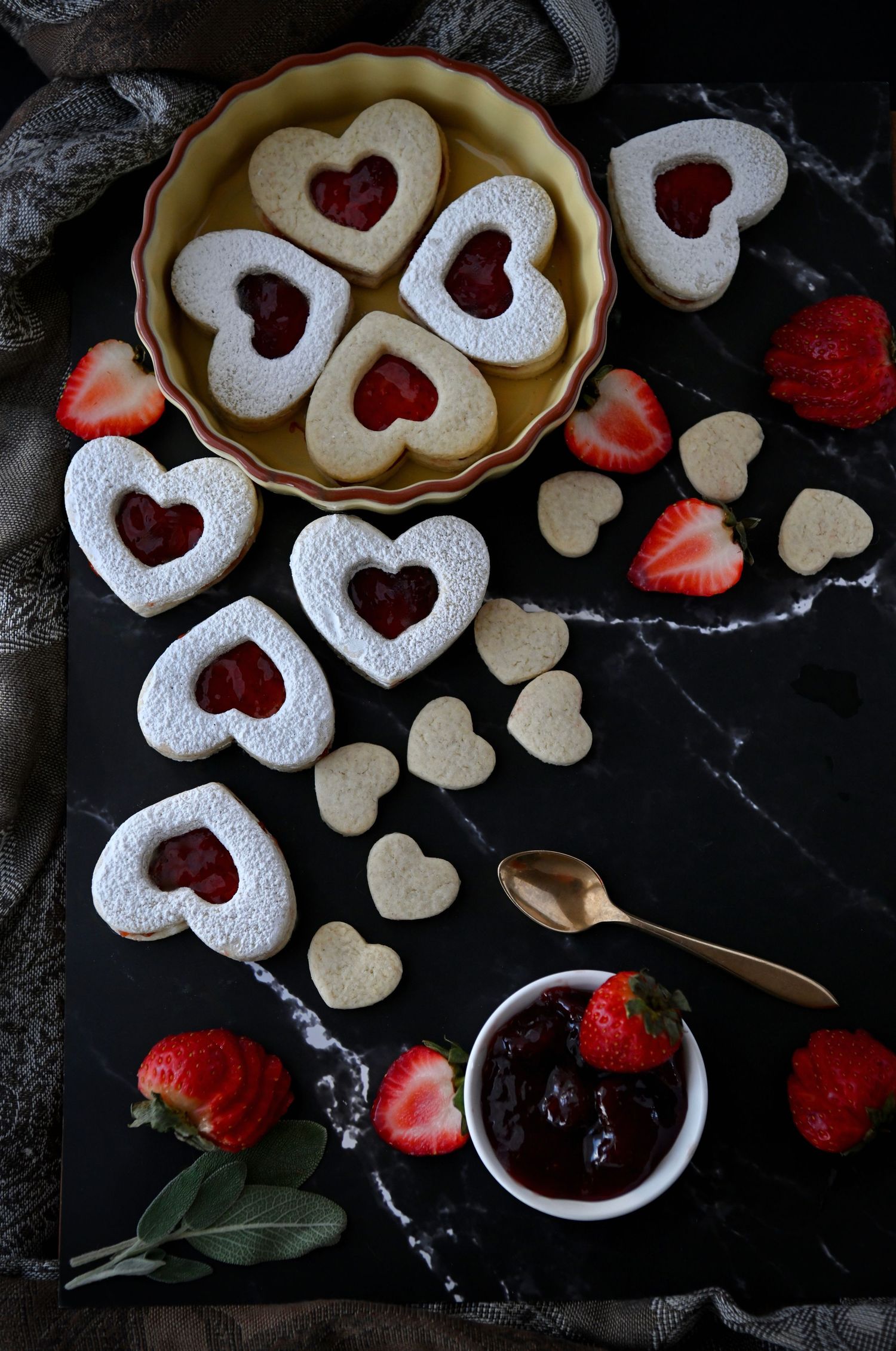 Heart-shaped linzer cookies with strawberry jam filling and powdered sugar arranged on dark marble surface with fresh berries.