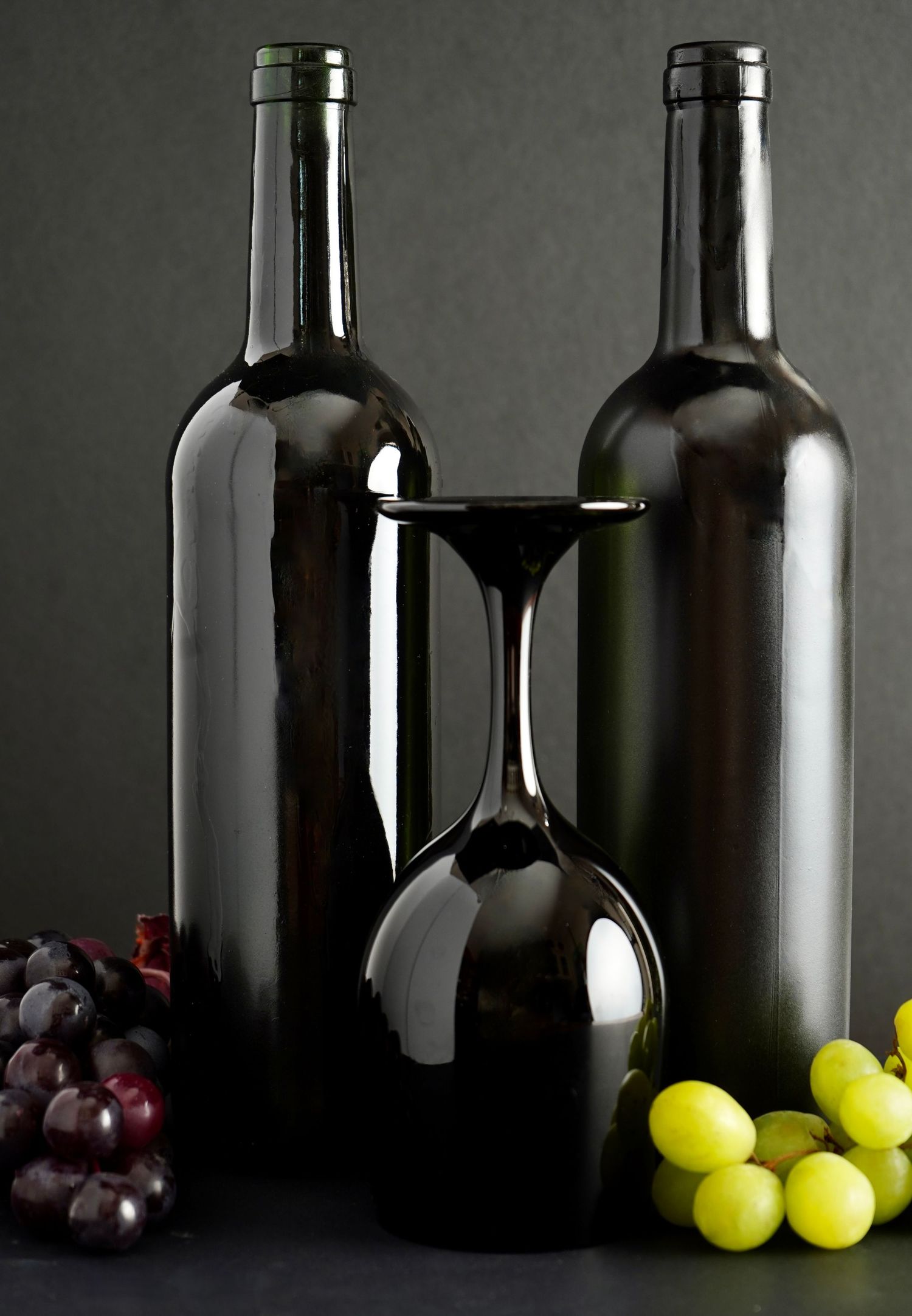 Still life with two dark wine bottles, an upside-down wine glass, and clusters of red and green grapes on dark background.