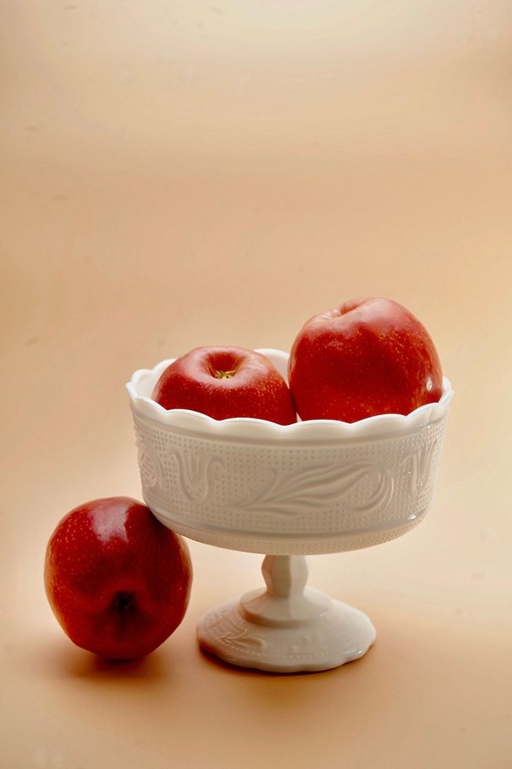 Red apples displayed next to an elegant white ceramic pedestal bowl on a beige background.
