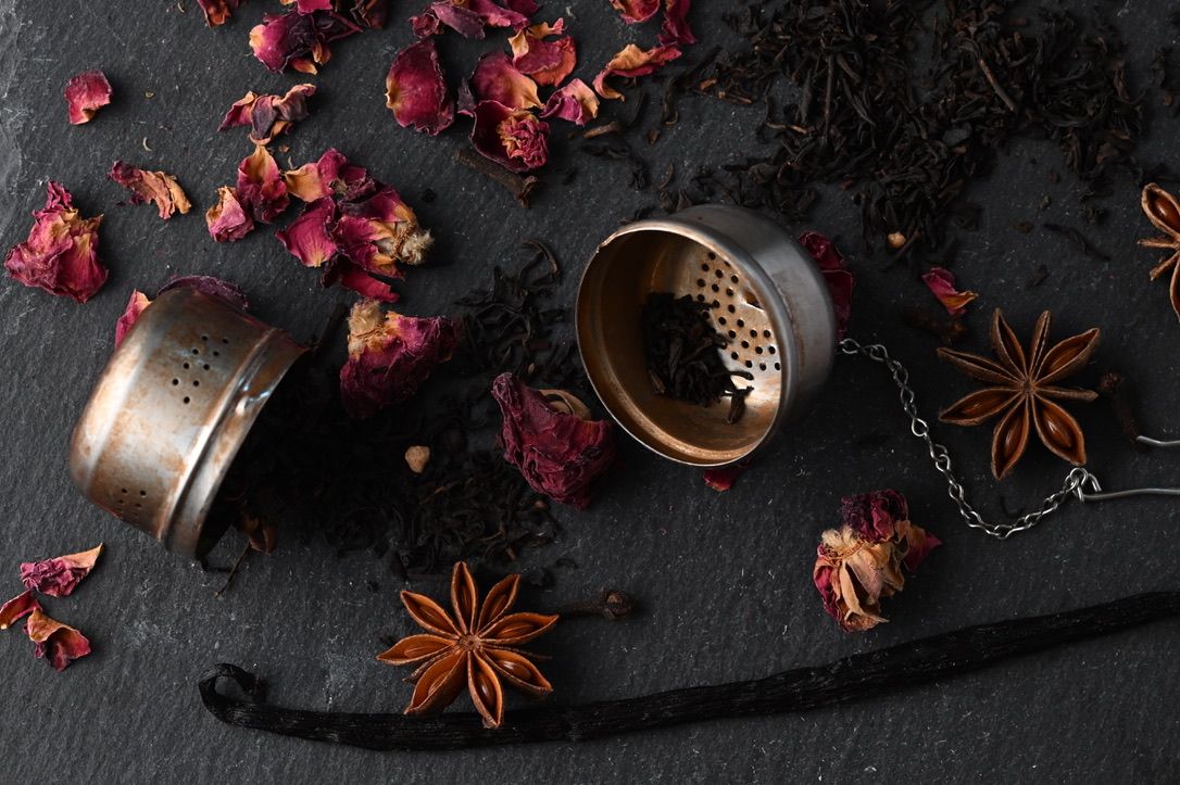 Star anise, black tea leaves and dried rose petals scattered around vintage tea egg on black surface.