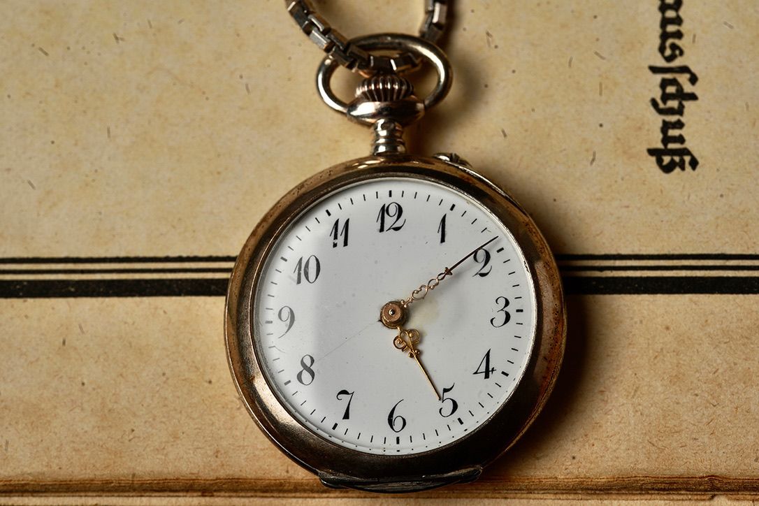 A vintage pocket watch with a white face lies on aged paper, casting a subtle shadow.