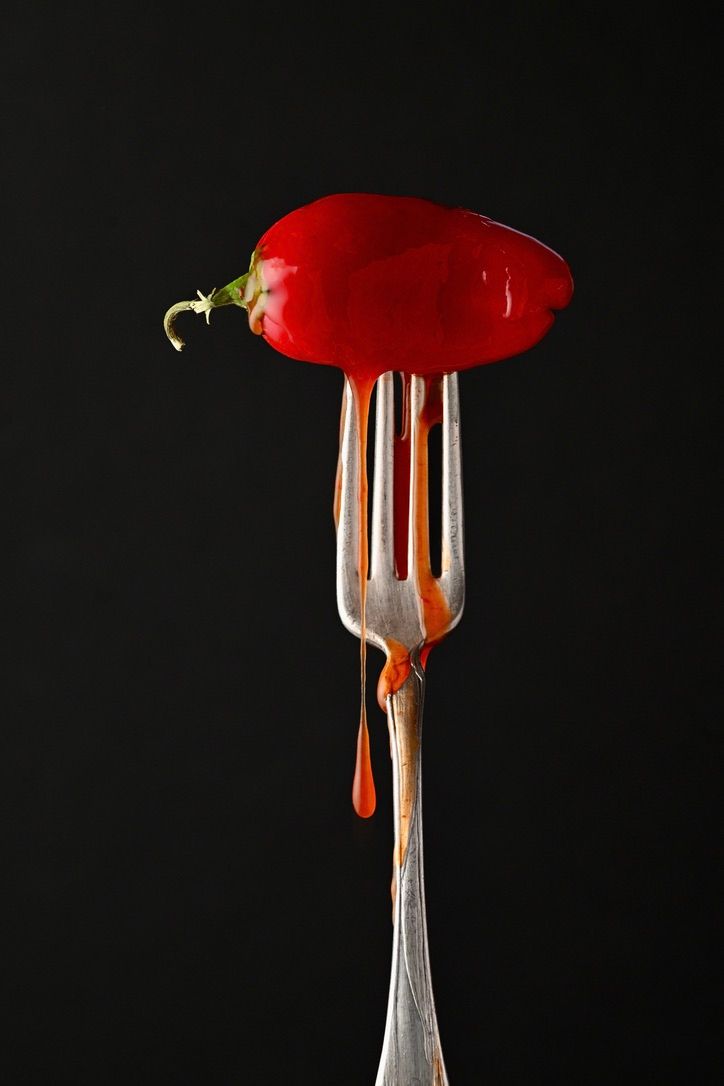 A bright red chili pepper impaled on a silver fork against a dark background shows a dramatic food photography composition.