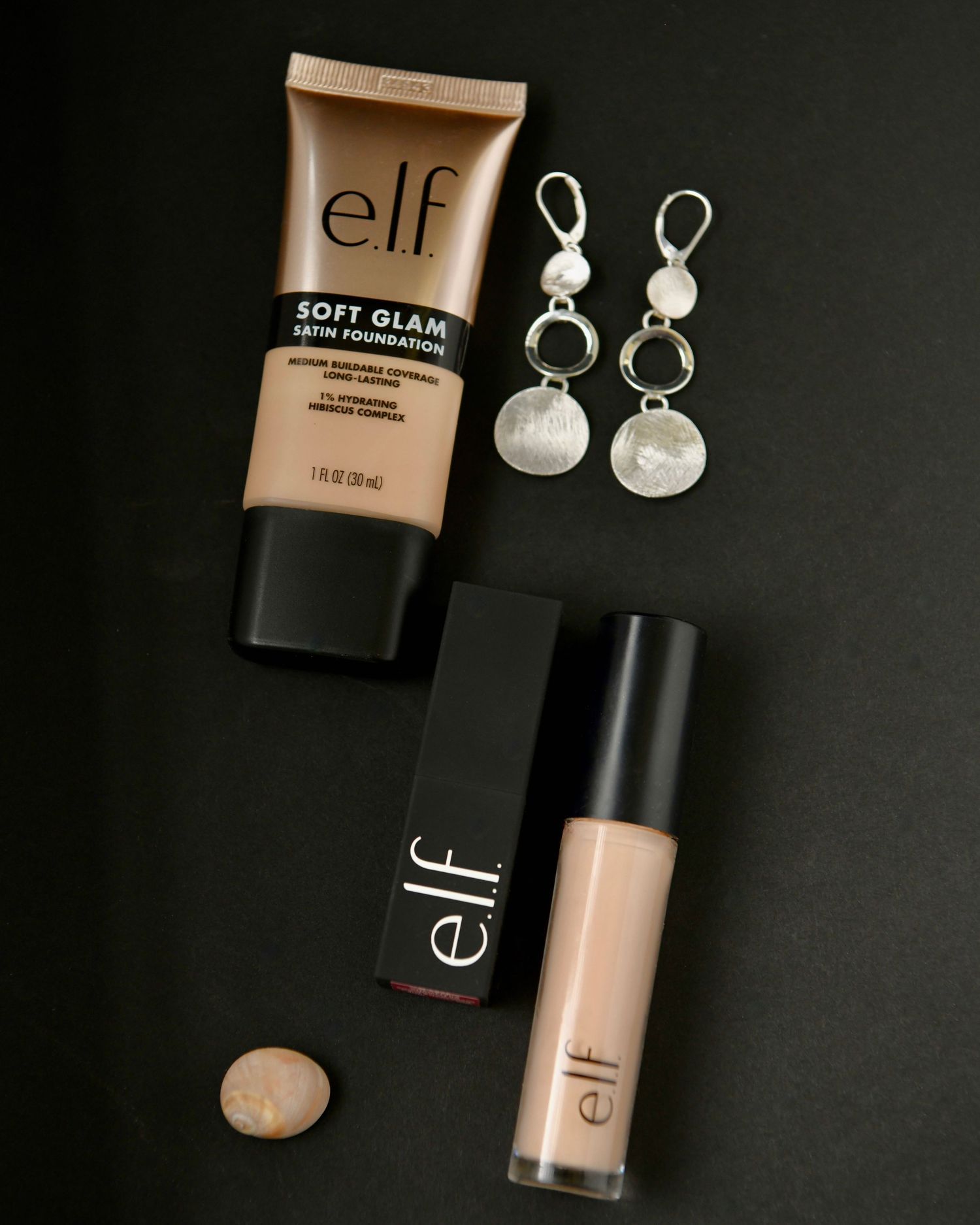 Makeup products from e.l.f. cosmetics including foundation and silver earrings arranged on dark background.