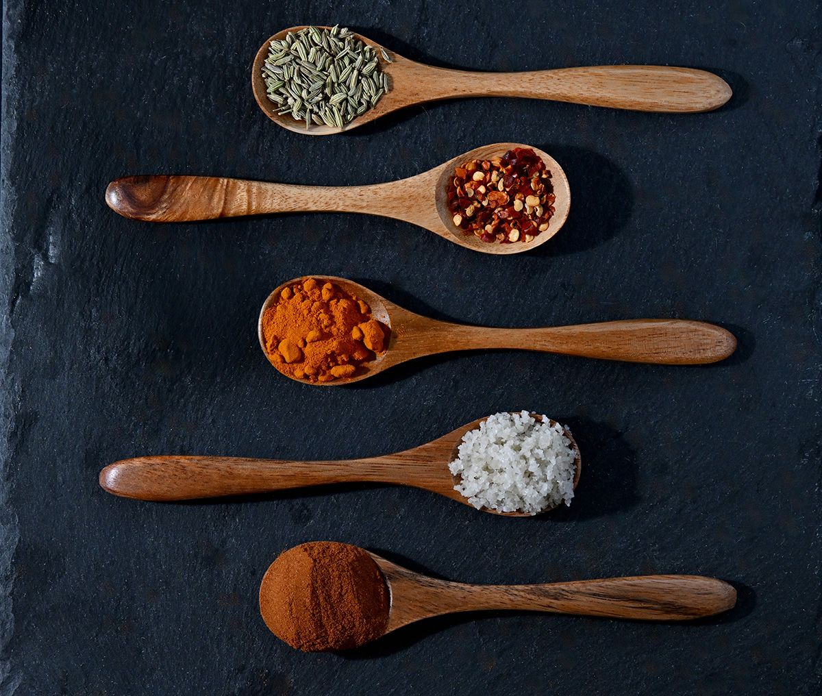 Wooden spoons filled with various colorful spices arranged on dark slate background.