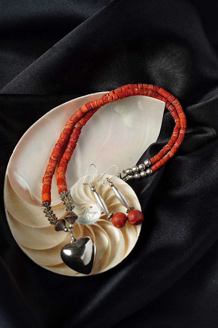 Coral bead necklace with silver heart pendant displayed on white nautilus shell against black fabric.