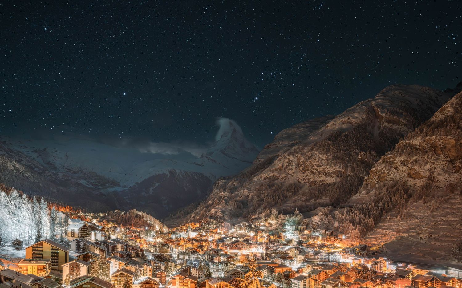Aerial view of illuminated mountain town at night with snow-covered peaks.