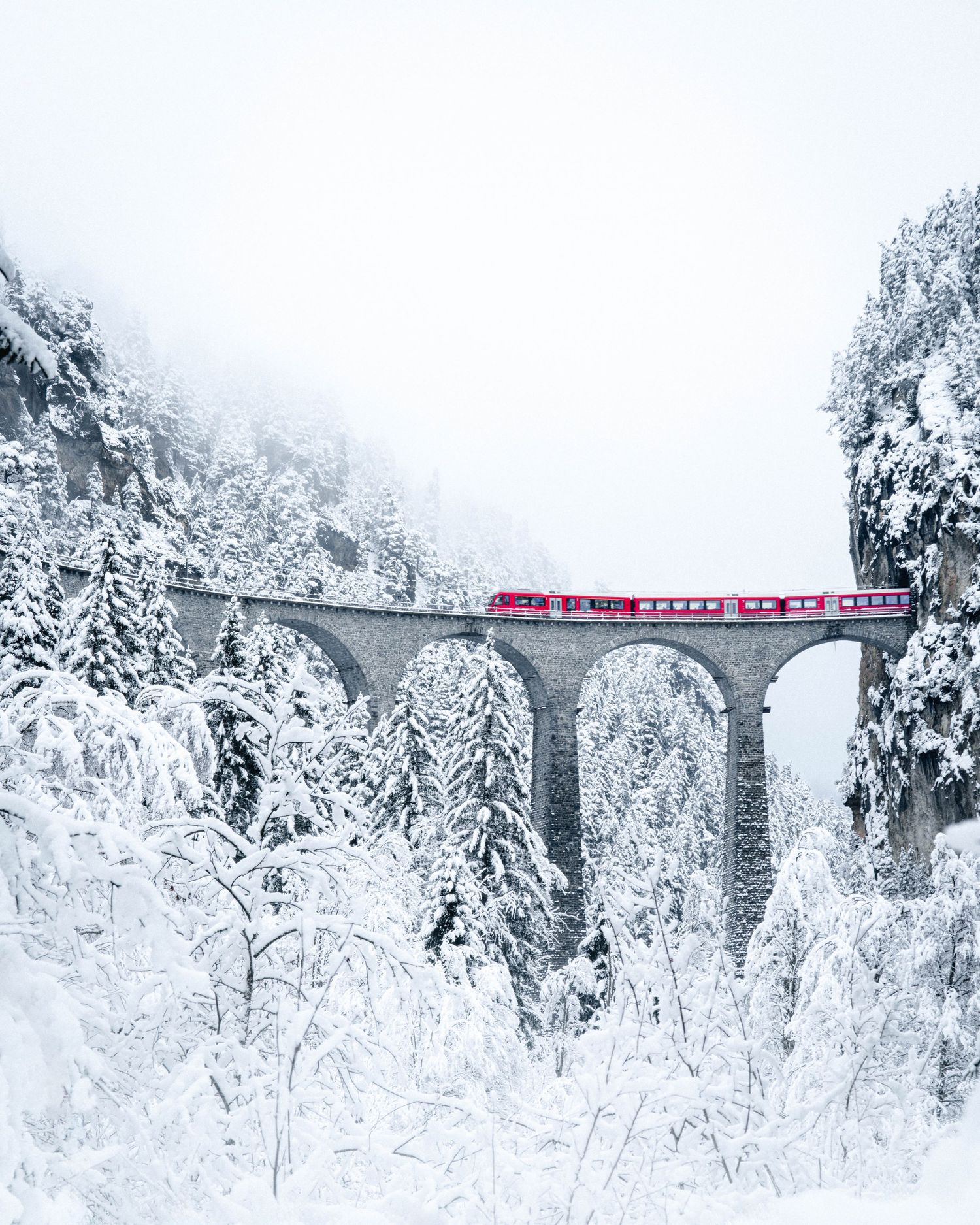 Red mountain train crossing a snow-covered viaduct bridge through a winter wonderland of frost-covered trees.