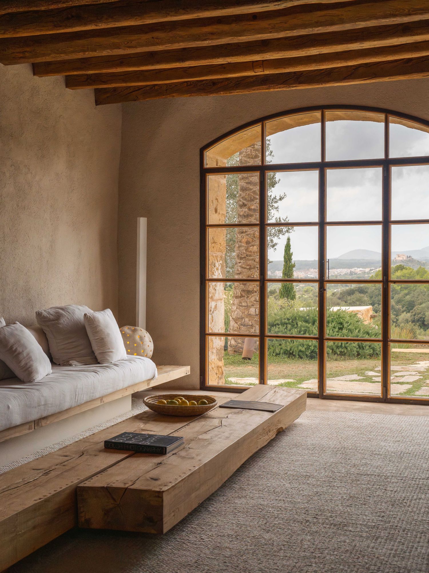 Minimalist meditation room with exposed wooden beams and large arched window overlooking countryside.