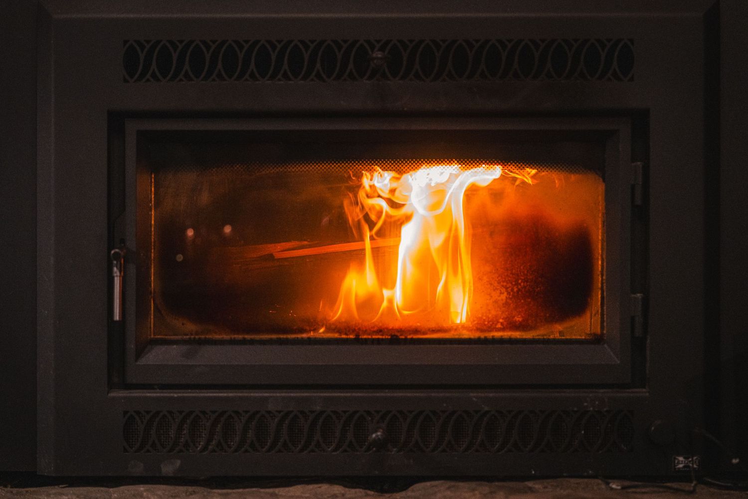 A glowing orange fire burns brightly behind the glass door of a cast iron wood stove with decorative details.