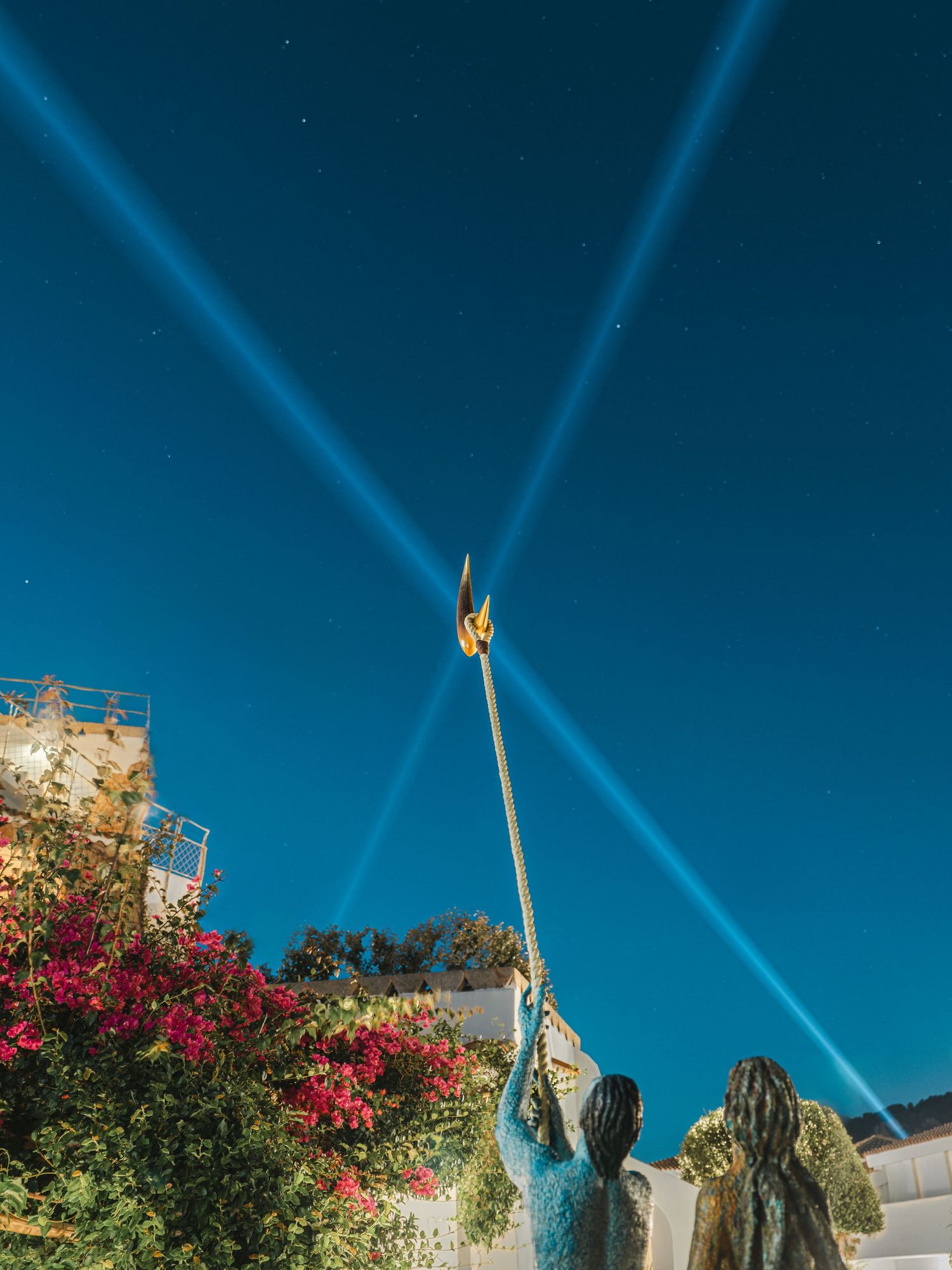 Dramatic blue spotlights crossing in night sky above pink bougainvillea flowers.