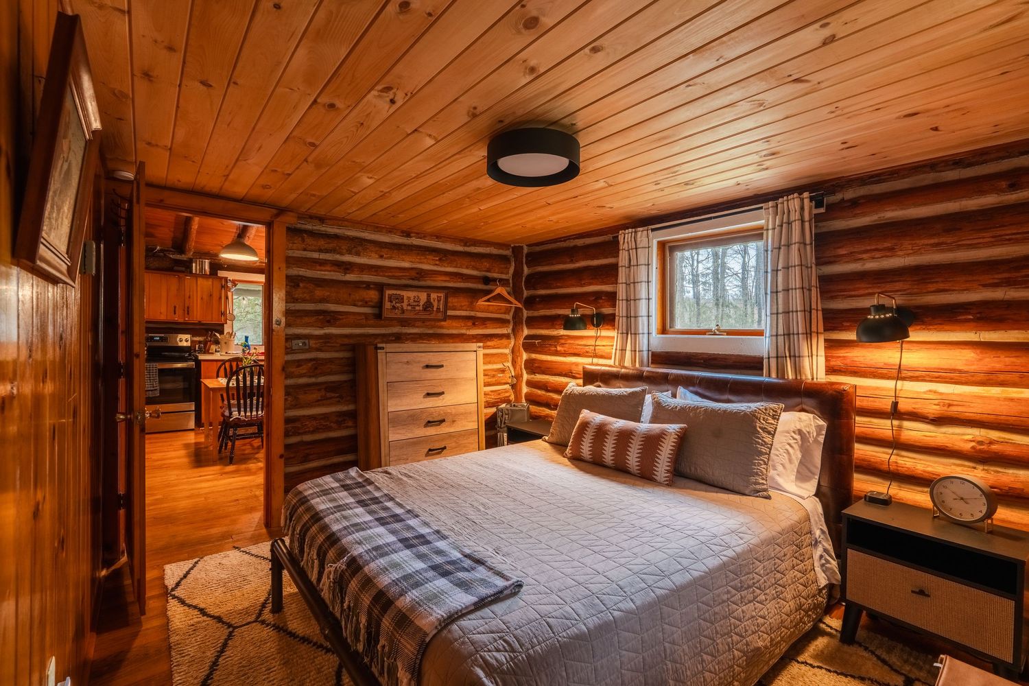 Cozy rustic bedroom interior with wooden log walls, warm lighting, and a plaid-accented bed in a cabin setting.