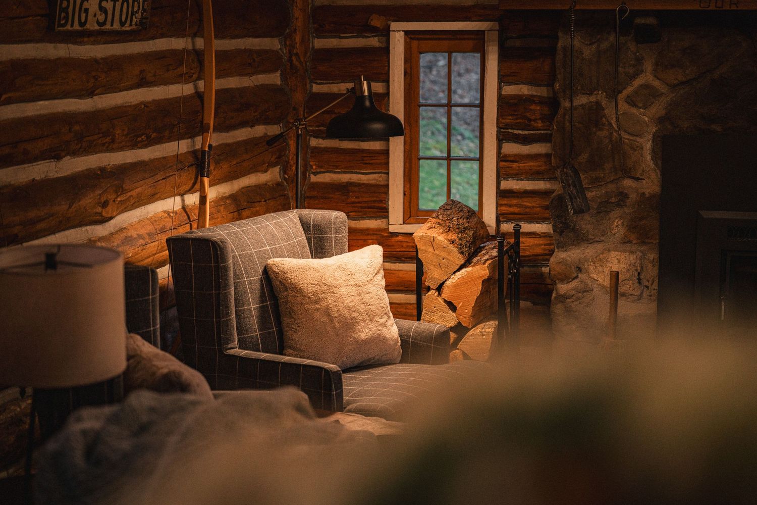 A cozy reading nook in a rustic log cabin features a comfortable armchair illuminated by warm lighting near a window.