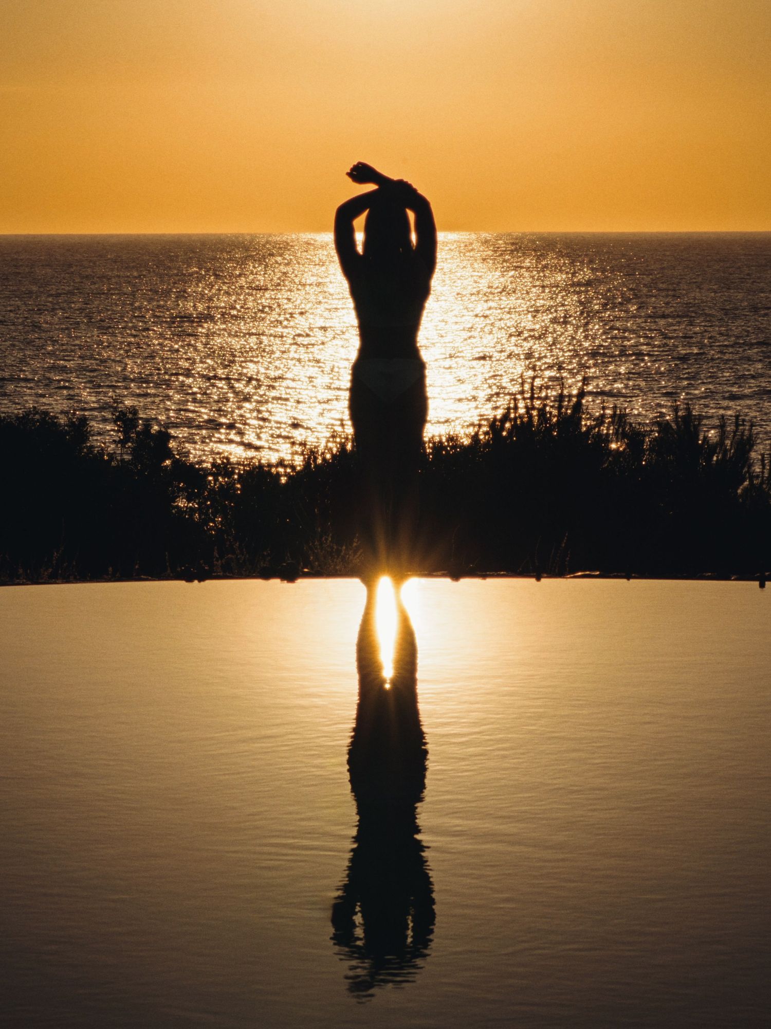 Silhouette of a person with arms raised reflected in an infinity pool at golden sunset over the ocean.